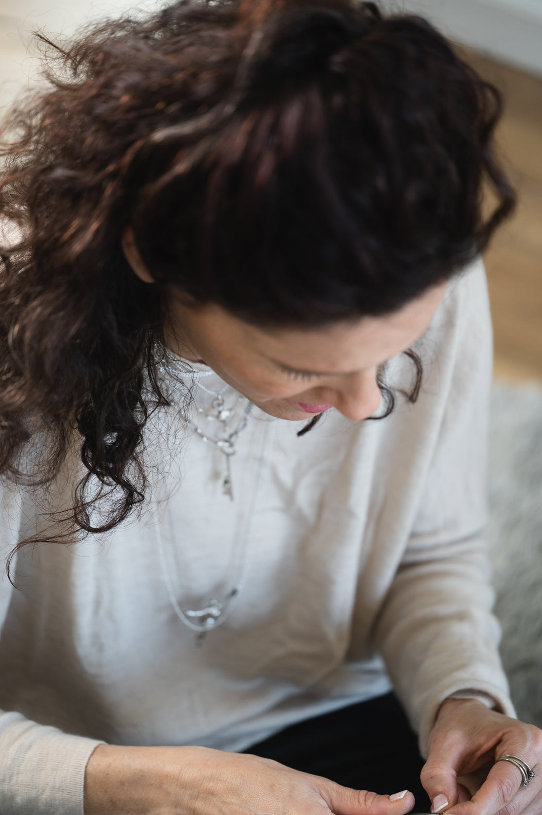 Woman with dark curly hair looking down at her hands, wearing layered silver necklaces and a silver ring