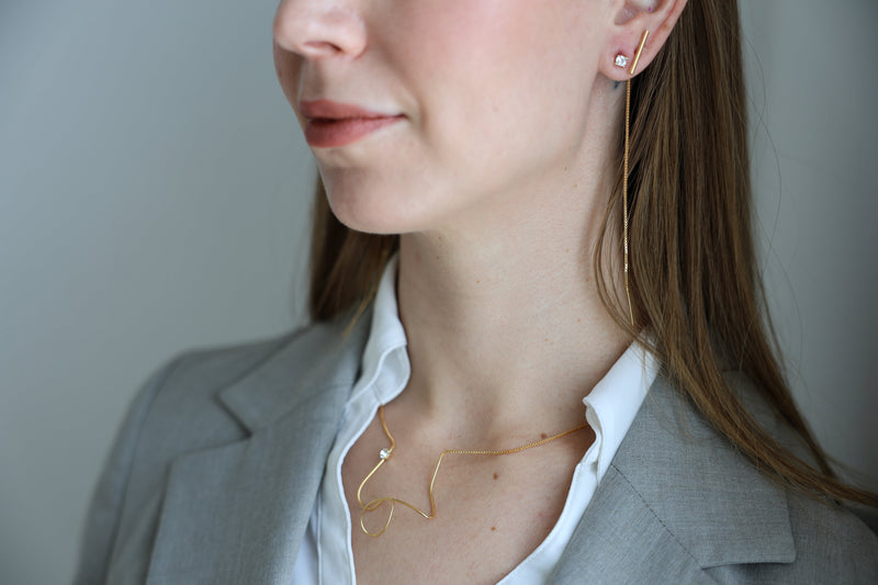 A close-up of a woman wearing a modern gold necklace with an abstract, freeform design and a small gemstone accent. She also wears a minimalist gold threader earring. Dressed in a gray blazer and white shirt, she showcases an elegant, contemporary style.